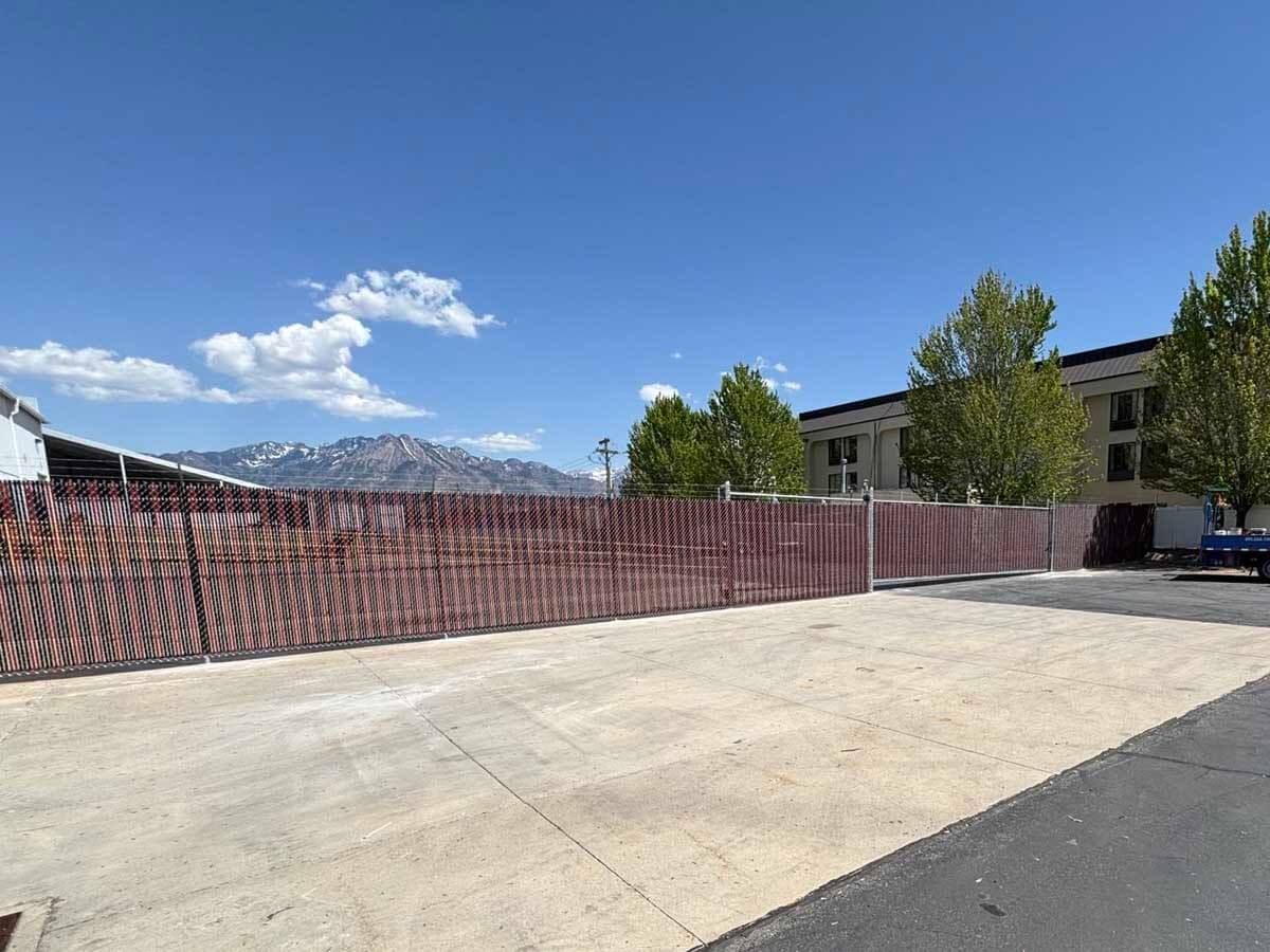 Empty paved lot beside a red construction fence, with trees and mountains under a blue sky.