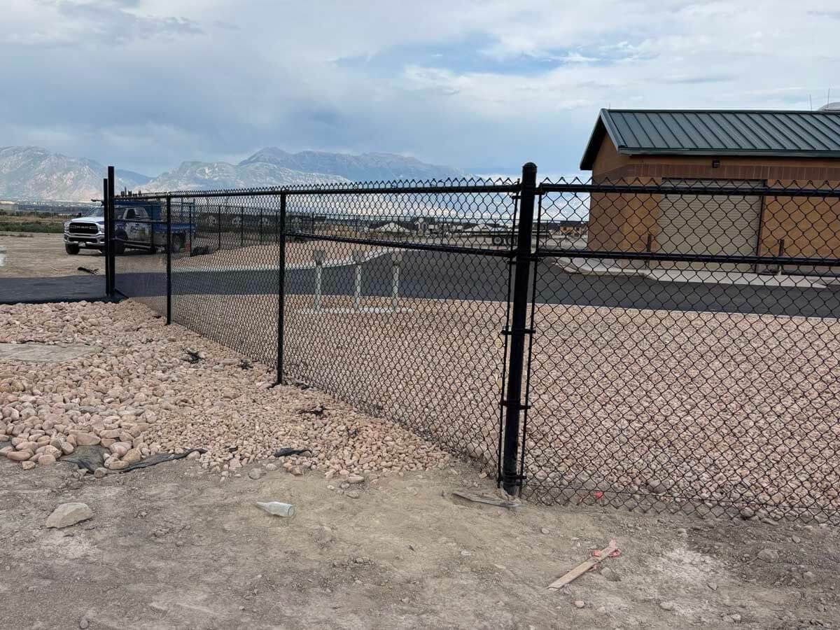 Chain-link fence beside a gravel lot, with a small shed and snowy mountains in the background.