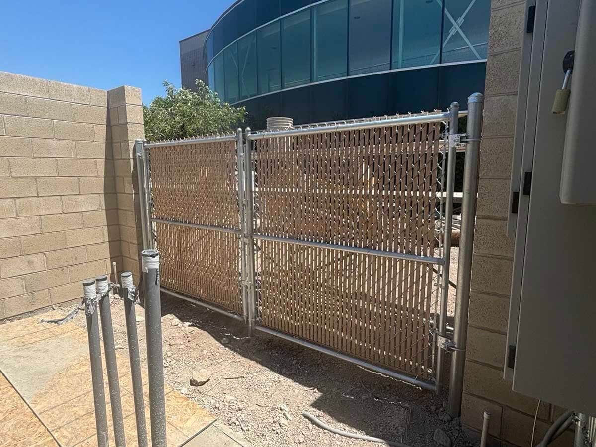 Metal gate beside a tan wall in a gravel yard, with a curved blue glass building in the background