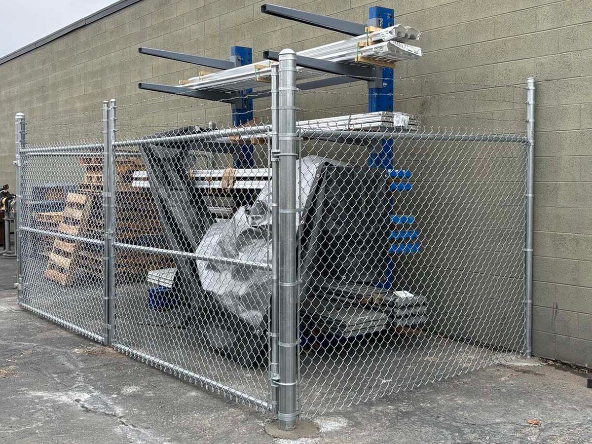 Chain-link fenced industrial machinery beside a brick building, with blue rollers and metal piping.