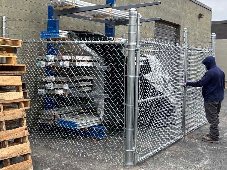 Worker opening a chain-link fence around stacked industrial equipment outside a warehouse