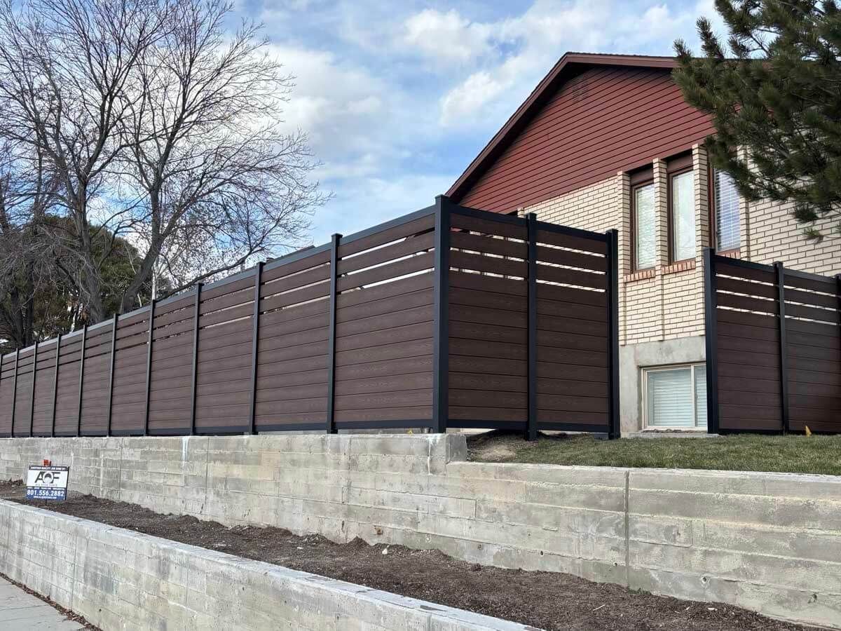 Long brown privacy fence on a concrete retaining wall beside a house and leafless trees