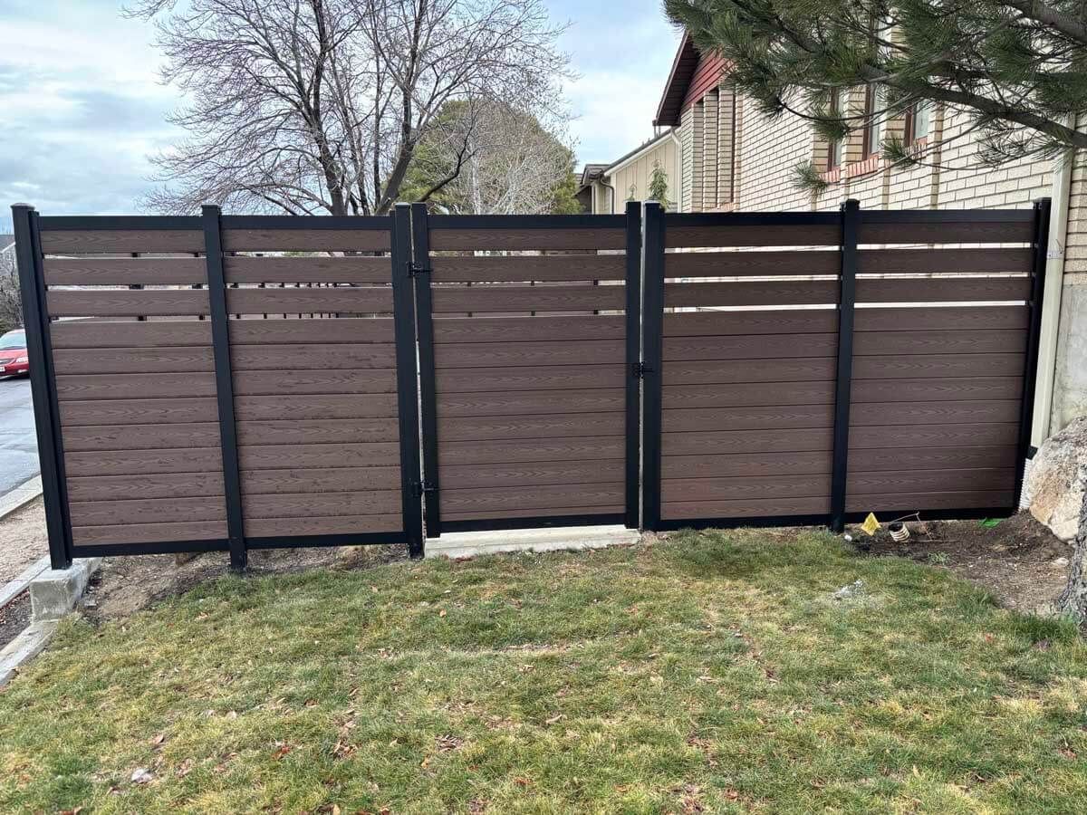Brown privacy fence with black posts and gate panels in a grassy yard beside a house