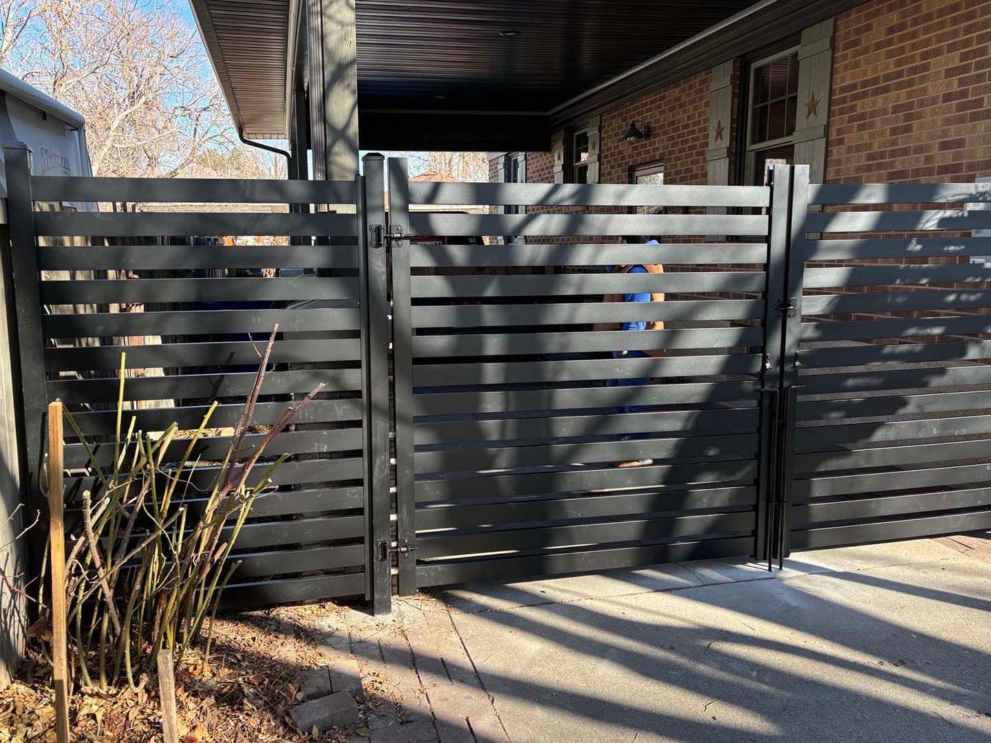 Black slatted metal gate under a carport, with sunlight and shadows across the sidewalk.