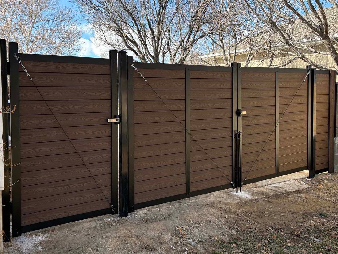 Brown wooden privacy fence with black metal posts along a gravel yard under bare trees