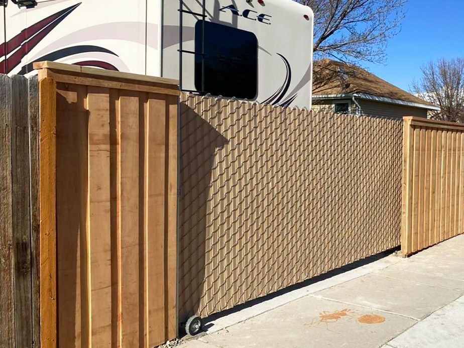 Chain-link and wooden fence along a driveway beside an RV in a sunny suburban yard.