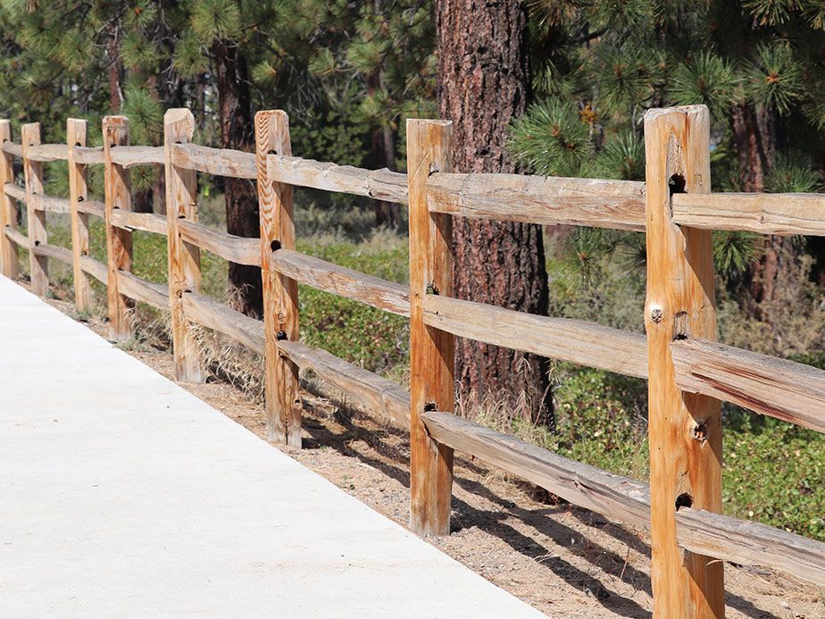 Wooden split-rail fence along a sunlit concrete path beside trees and brush