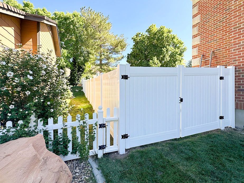 White fence and gate beside a brick house, with green lawn, bushes, and trees in a sunny yard
