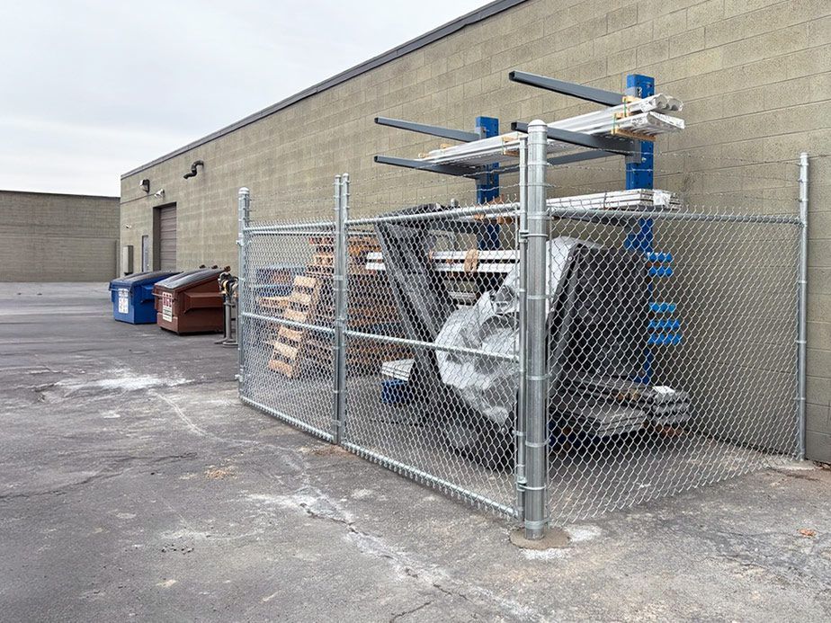 Chain-link fenced storage area beside a beige warehouse, filled with ladders, pallets, and metal racks.