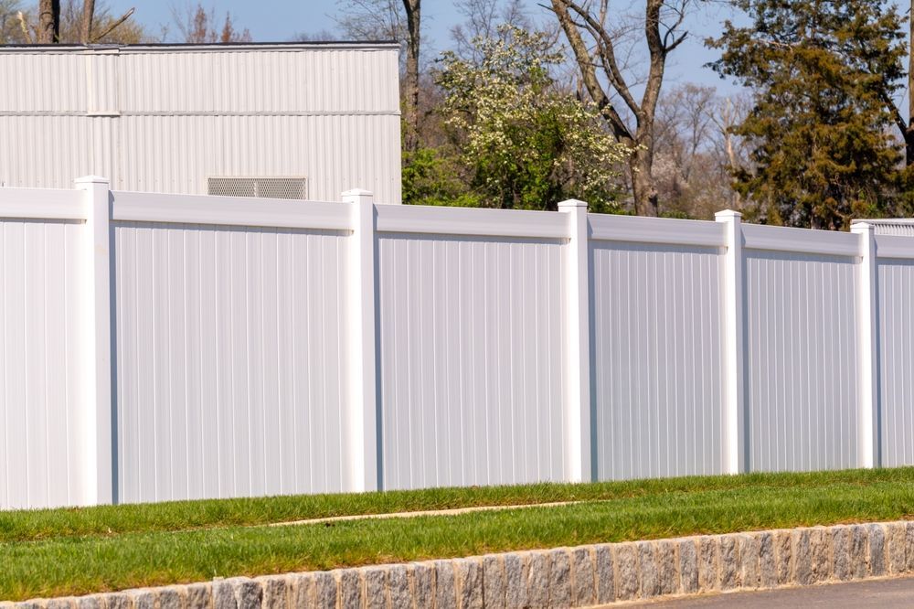 White vinyl privacy fence along a grassy roadside with a building and trees behind it