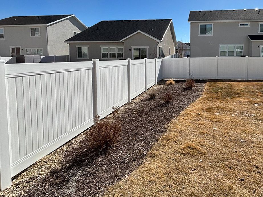 Backyard with white vinyl fence, dry grass, and mulched planting strip in a suburban neighborhood