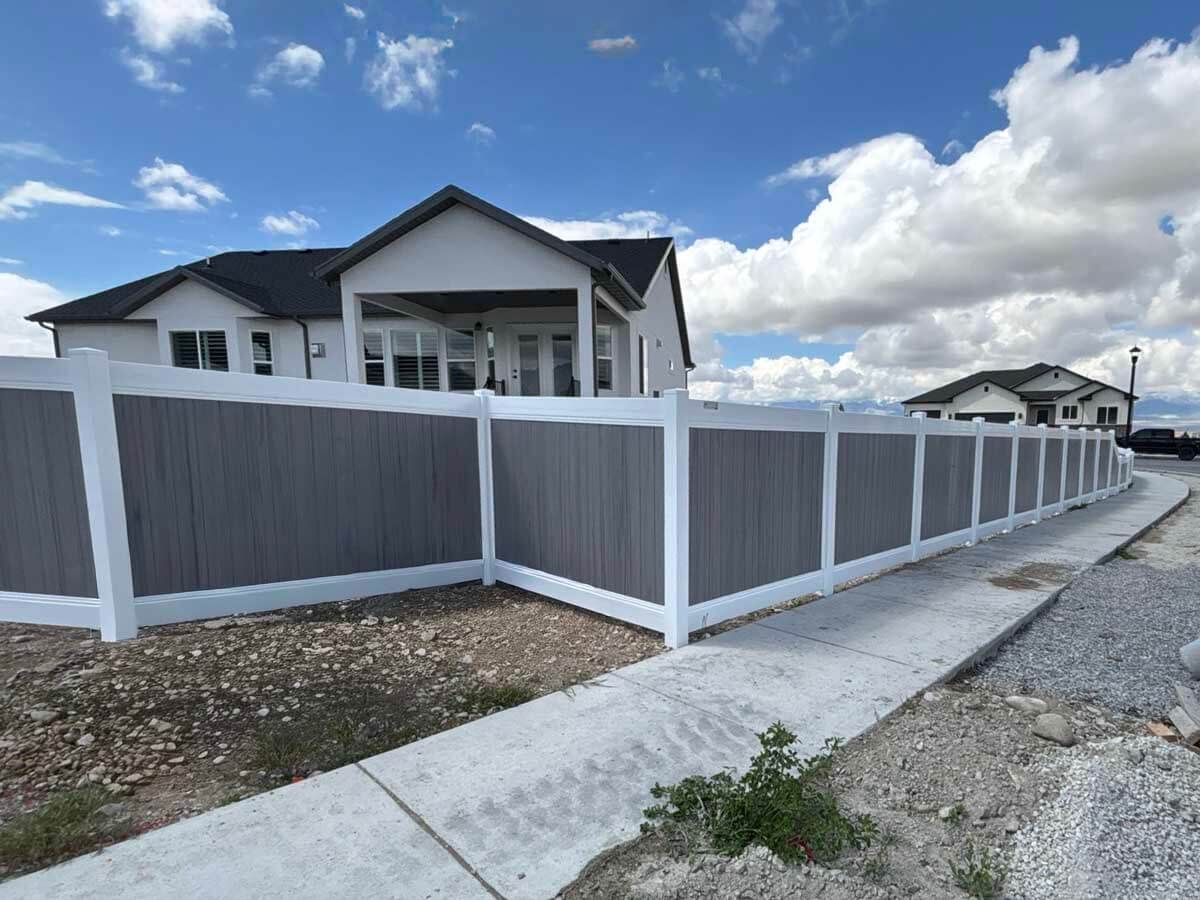 Gray-and-white house behind a tall gray fence along a gravel roadside under a cloudy sky