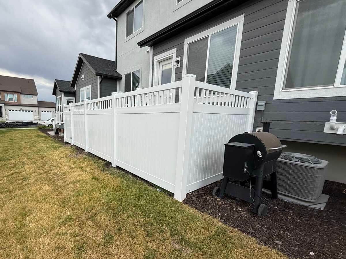 White deck and railing beside a gray house, with a black grill on a small landscaped strip.