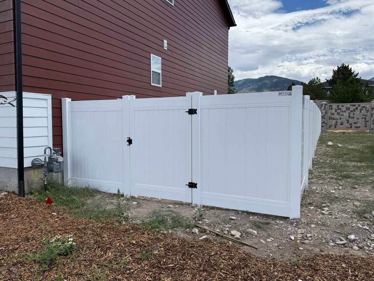 White double gate beside a red-sided house, opening to a gravel driveway and yard