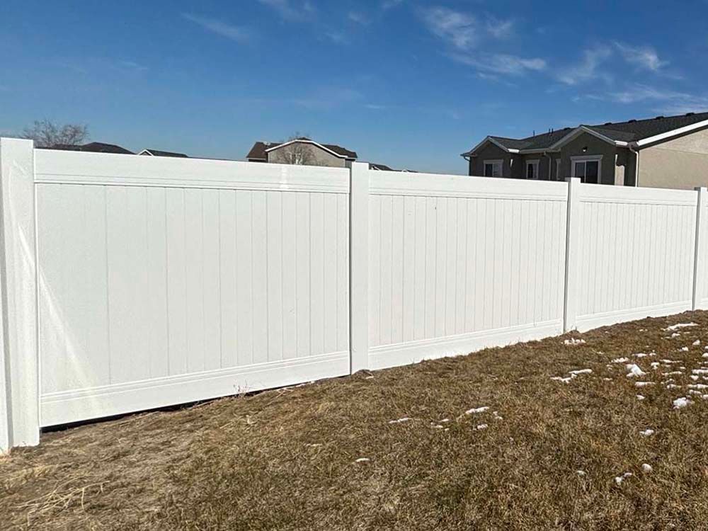 White vinyl fence along a sloped brown yard under a blue sky, with houses in the background