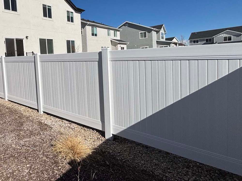 White vinyl privacy fence beside beige townhouses under a clear blue sky, with a shadowed gravel yard