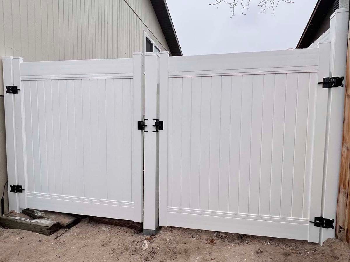 White wooden double gate with black hinges and latches in a fenced outdoor driveway.