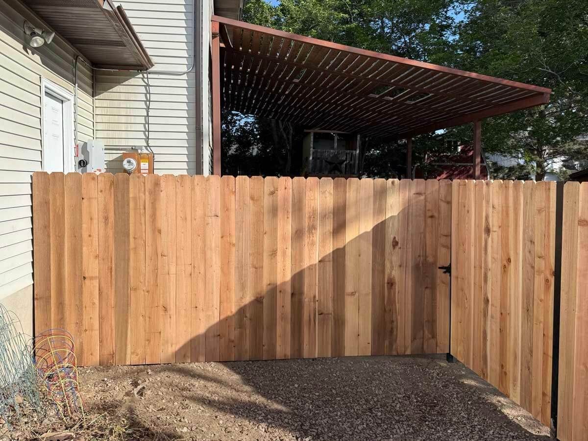 Wooden privacy fence beside a house and gravel driveway, with a covered patio in the background.