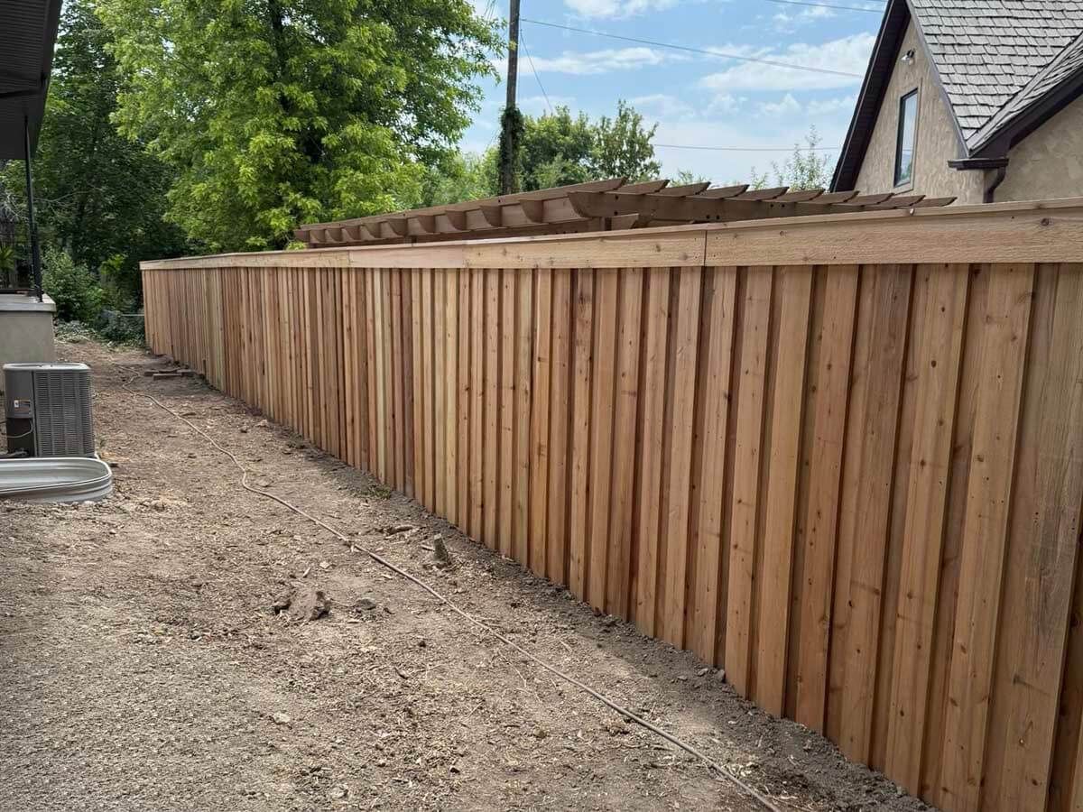 Long wooden fence along a dirt path beside a house and trees under a partly cloudy sky