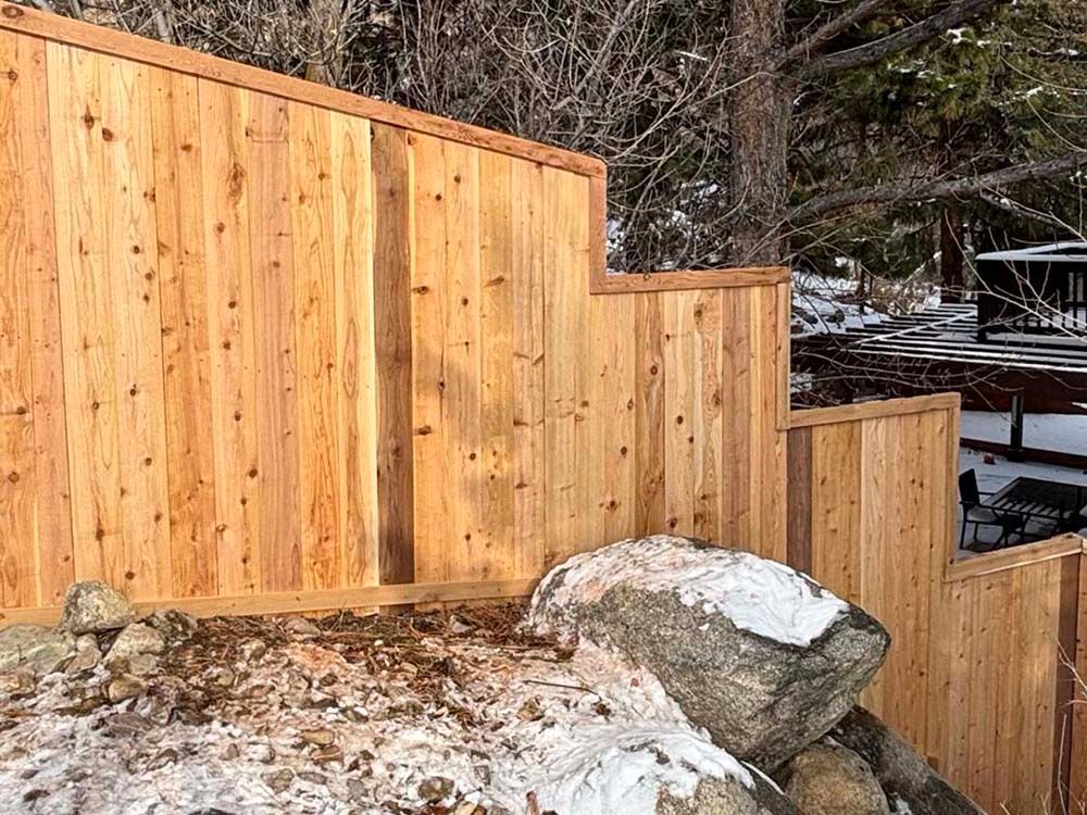 Snow-dusted wooden fence and steps beside a large rock in a wooded yard