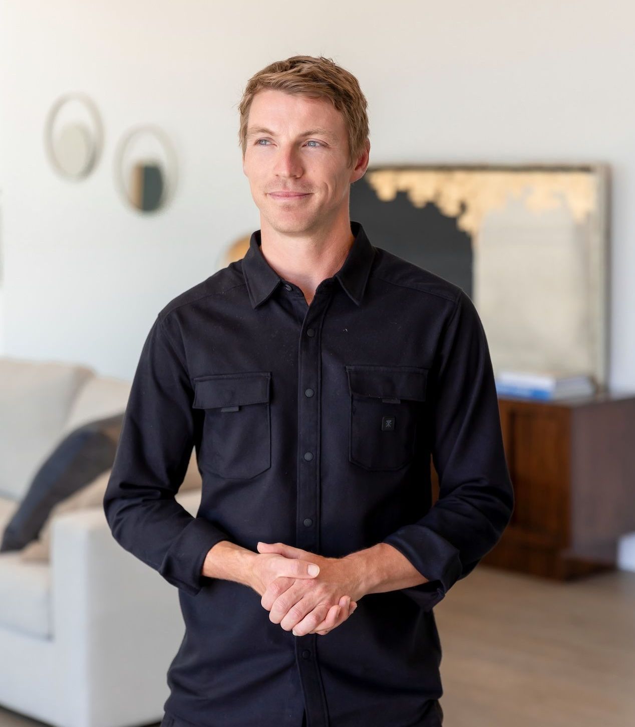 A man in a black shirt is standing in a living room with his hands folded