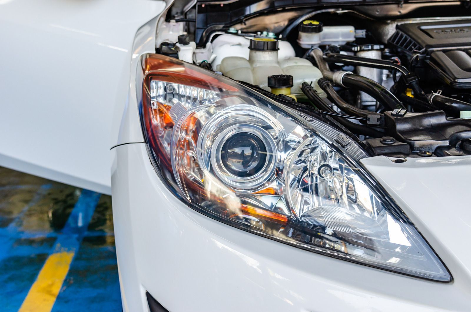 White car headlight and open hood, engine visible, garage setting.