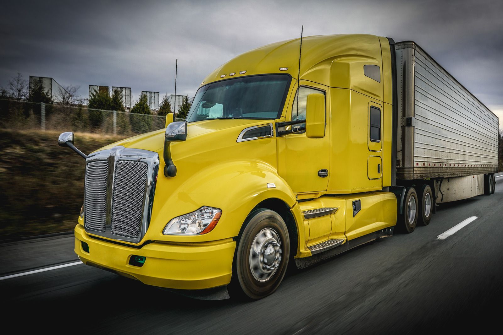 Yellow semi-truck with trailer driving on a highway. Overcast sky.