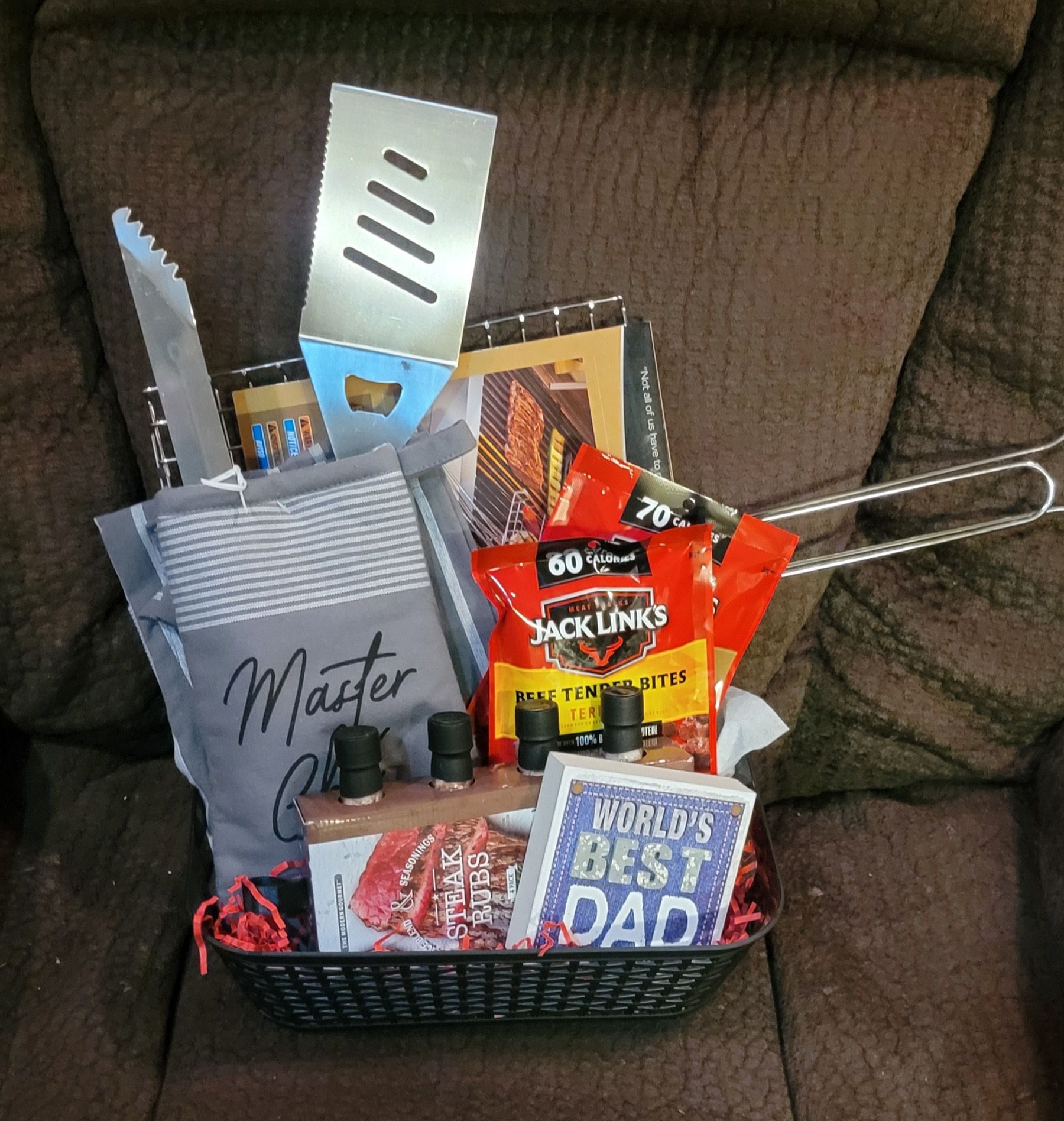Gift basket with grilling tools, snacks, and a “World’s Best Dad” card on a sofa.