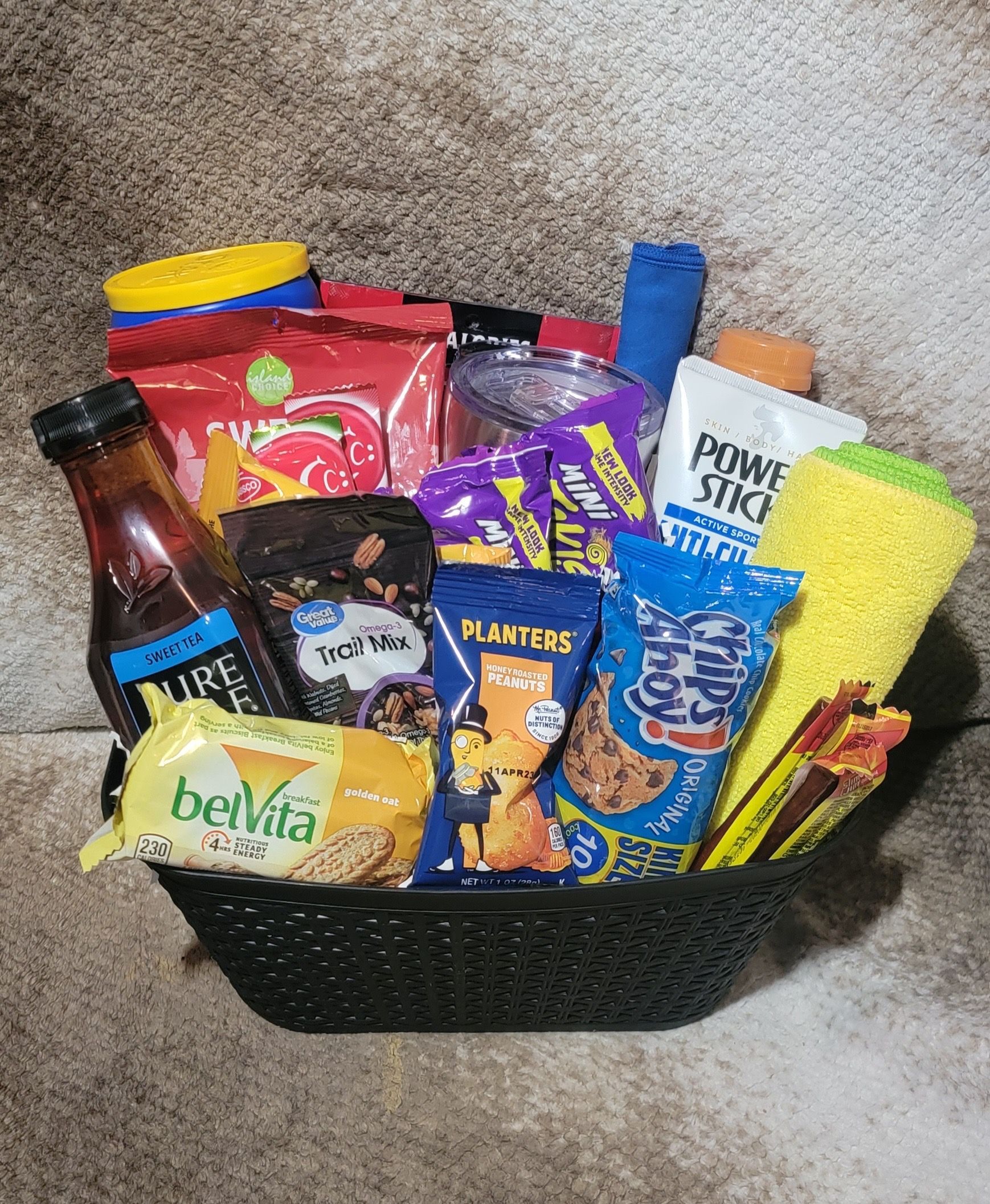 Black basket filled with assorted snacks, candy, and a yellow sponge on a carpet.
