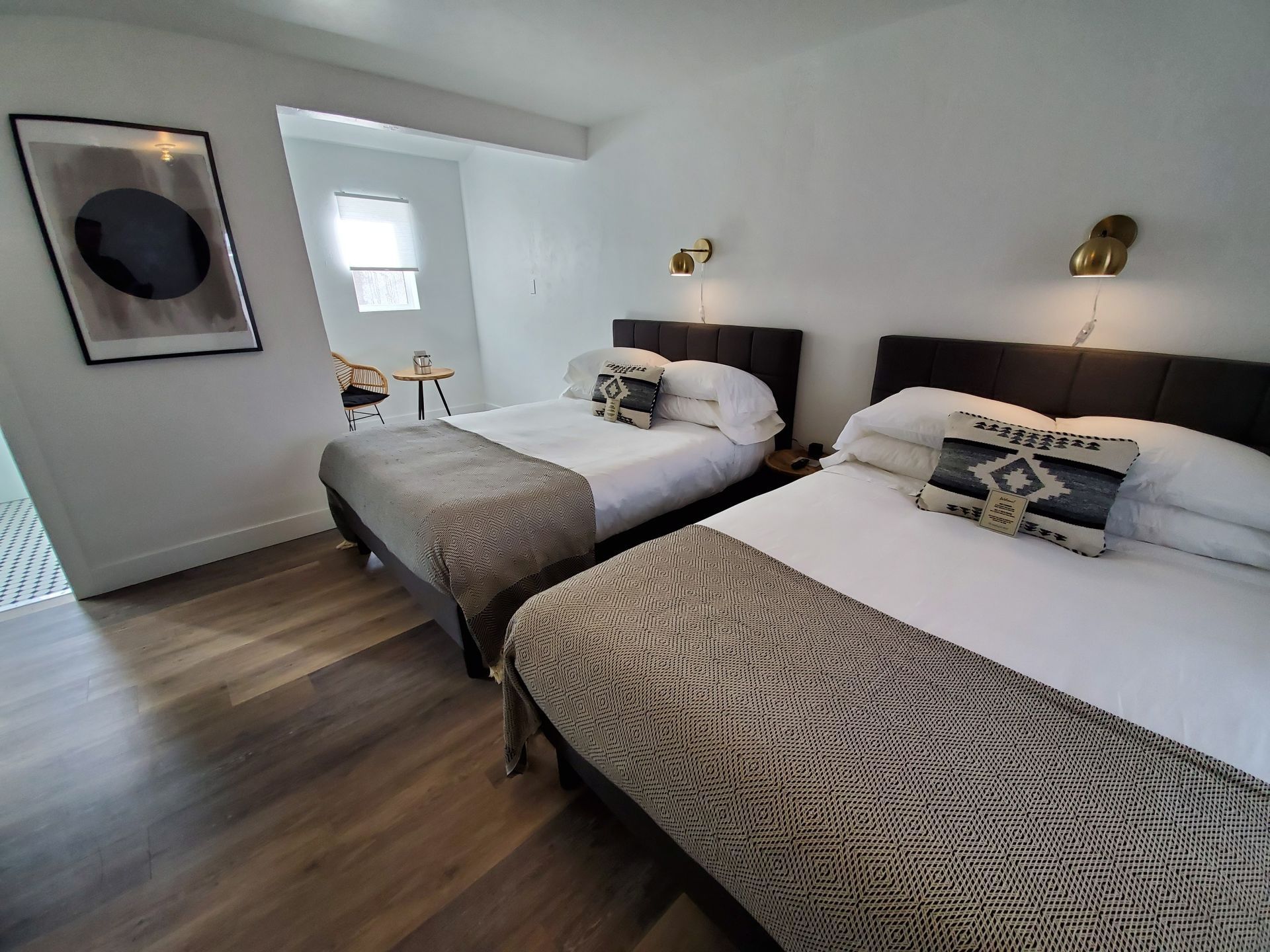 Two beds in a white room. Dark brown headboards, gold sconces, art on the wall, and wood flooring.