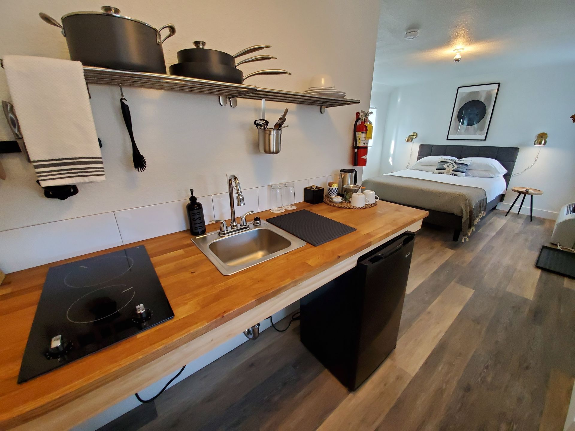Small kitchen in a studio apartment with a sink, stove, and shelves above. A bed is in the background.