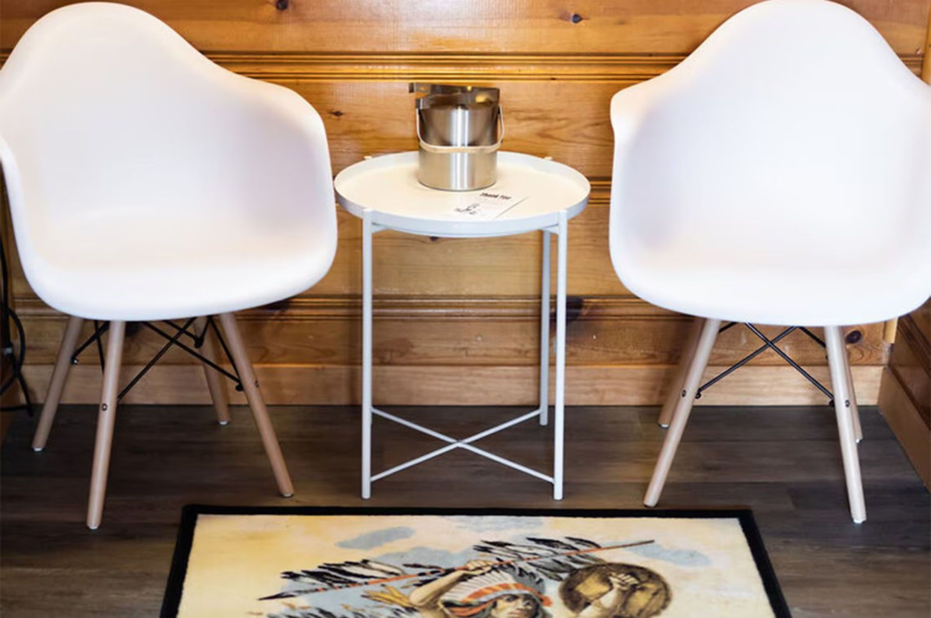 Two white chairs flank a small round table with a gold coffee maker. Wood paneled wall and floor.