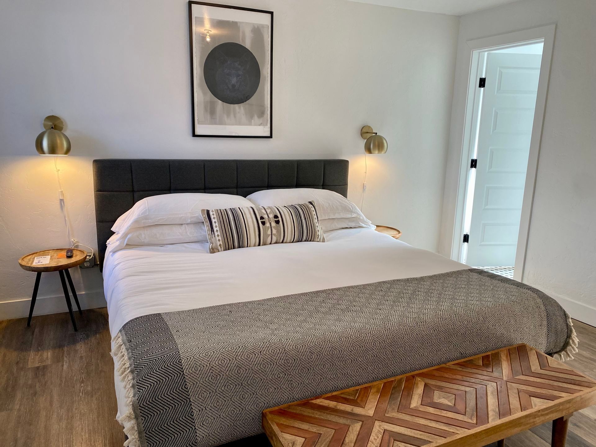 Bedroom with a large bed, decorative pillows, art, and a bench. Light walls, gold sconces, and a view into the bathroom.