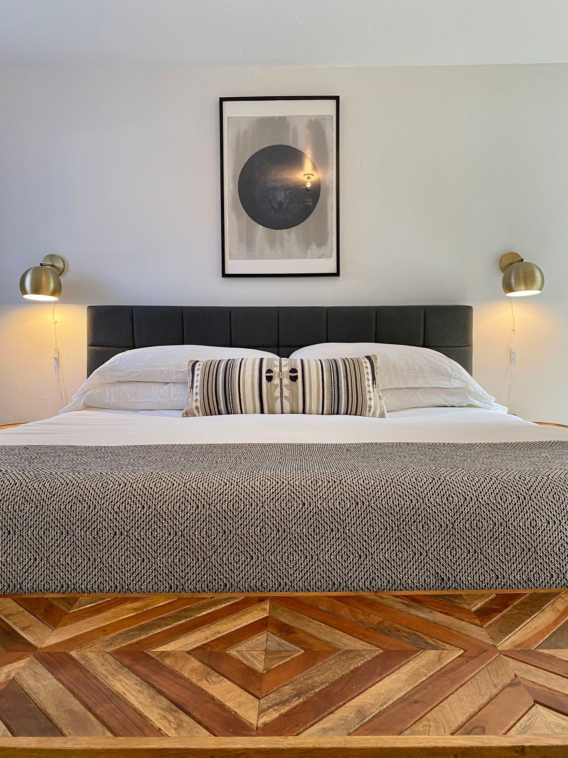 Bedroom with a dark gray headboard, art, decorative pillows, patterned blanket, and wall sconces.