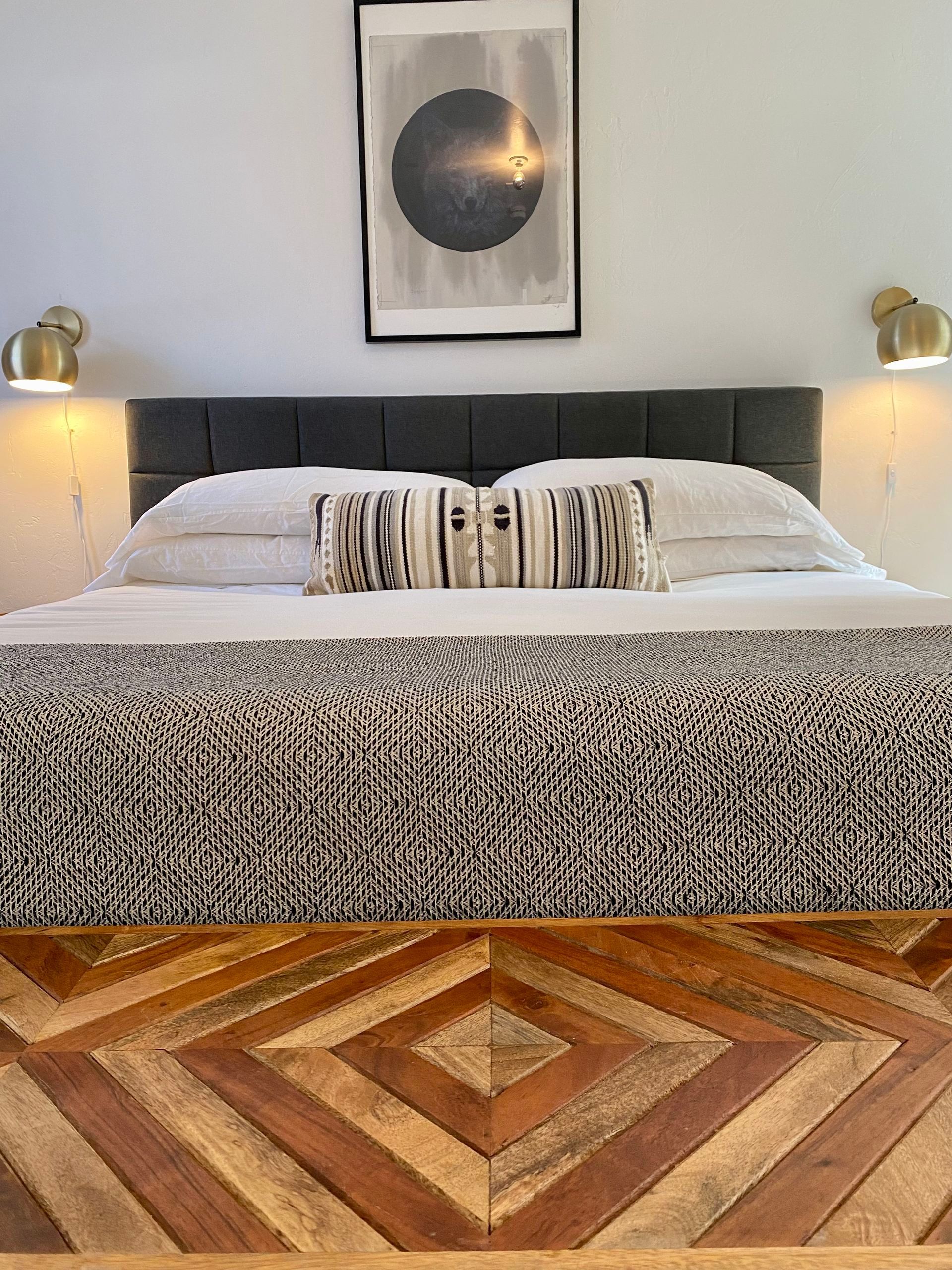 Bedroom with bed: gray headboard, geometric wood footboard, white bedding, black & white blanket, art, and wall sconces.
