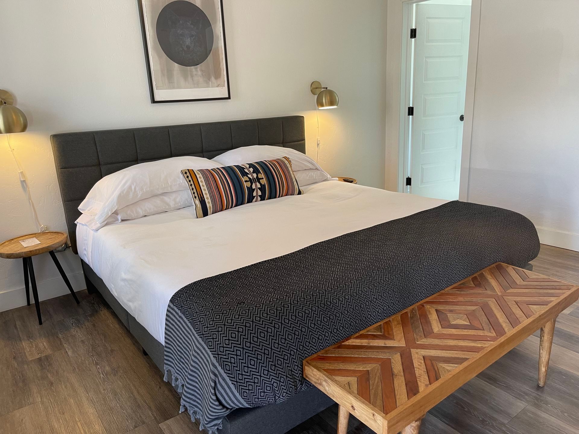 Bedroom with king-size bed, bench, side table, artwork, and wall sconces. Neutral colors.
