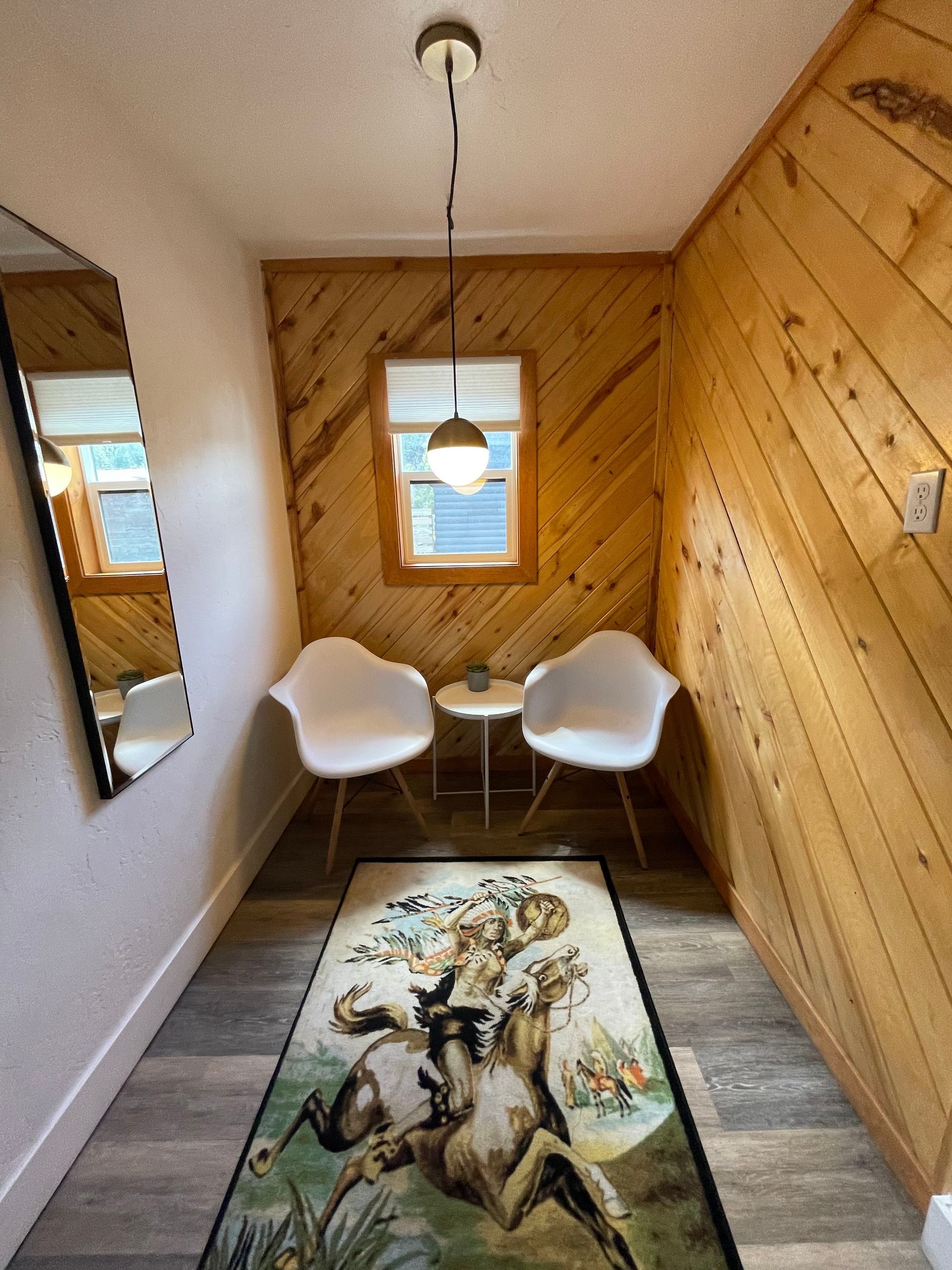 Narrow room with white chairs, small table, rug, wood paneling, mirror, and pendant light.