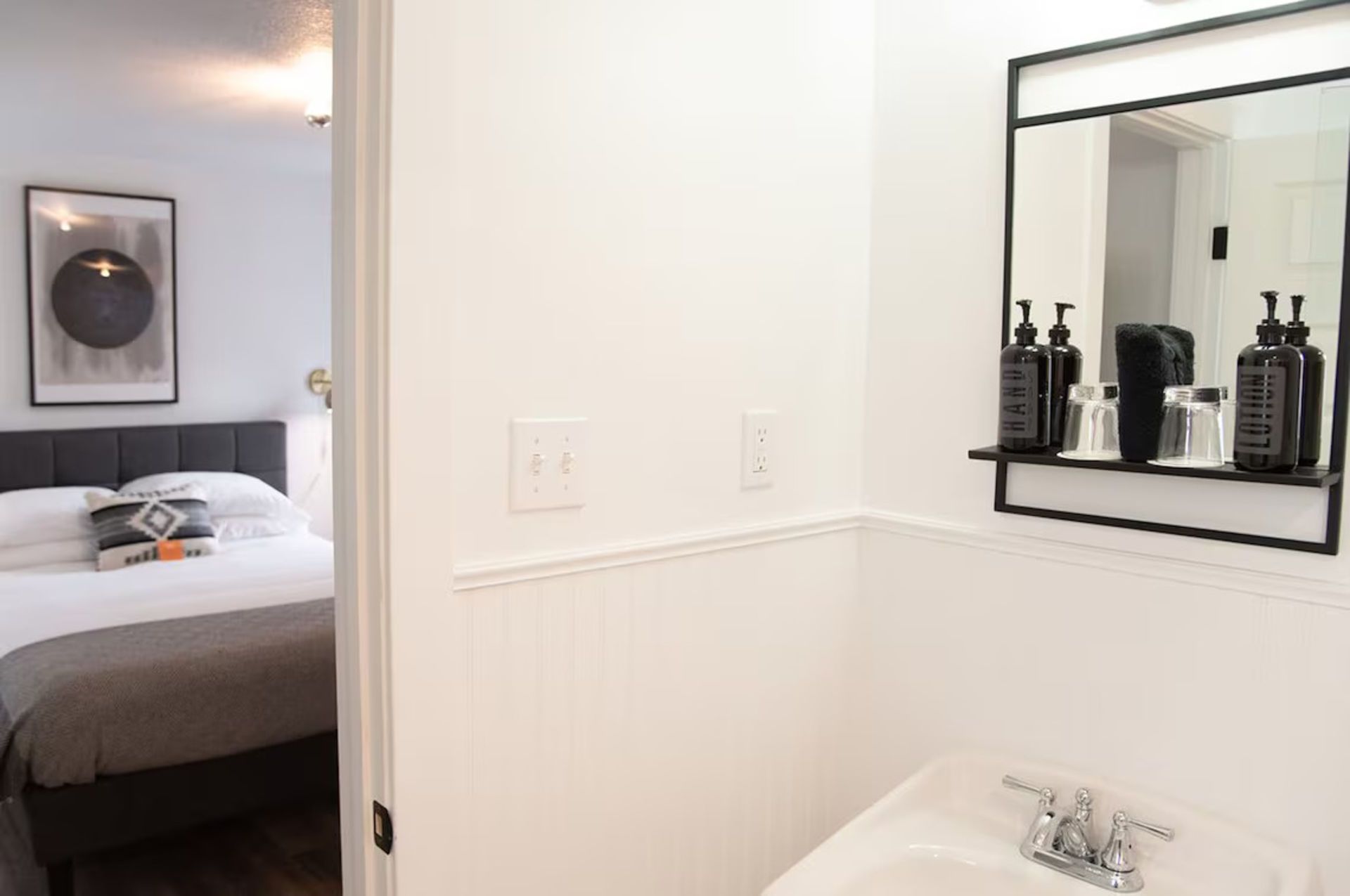 Bathroom doorway with a bedroom in view; white walls and sink, black framed mirror with toiletries.