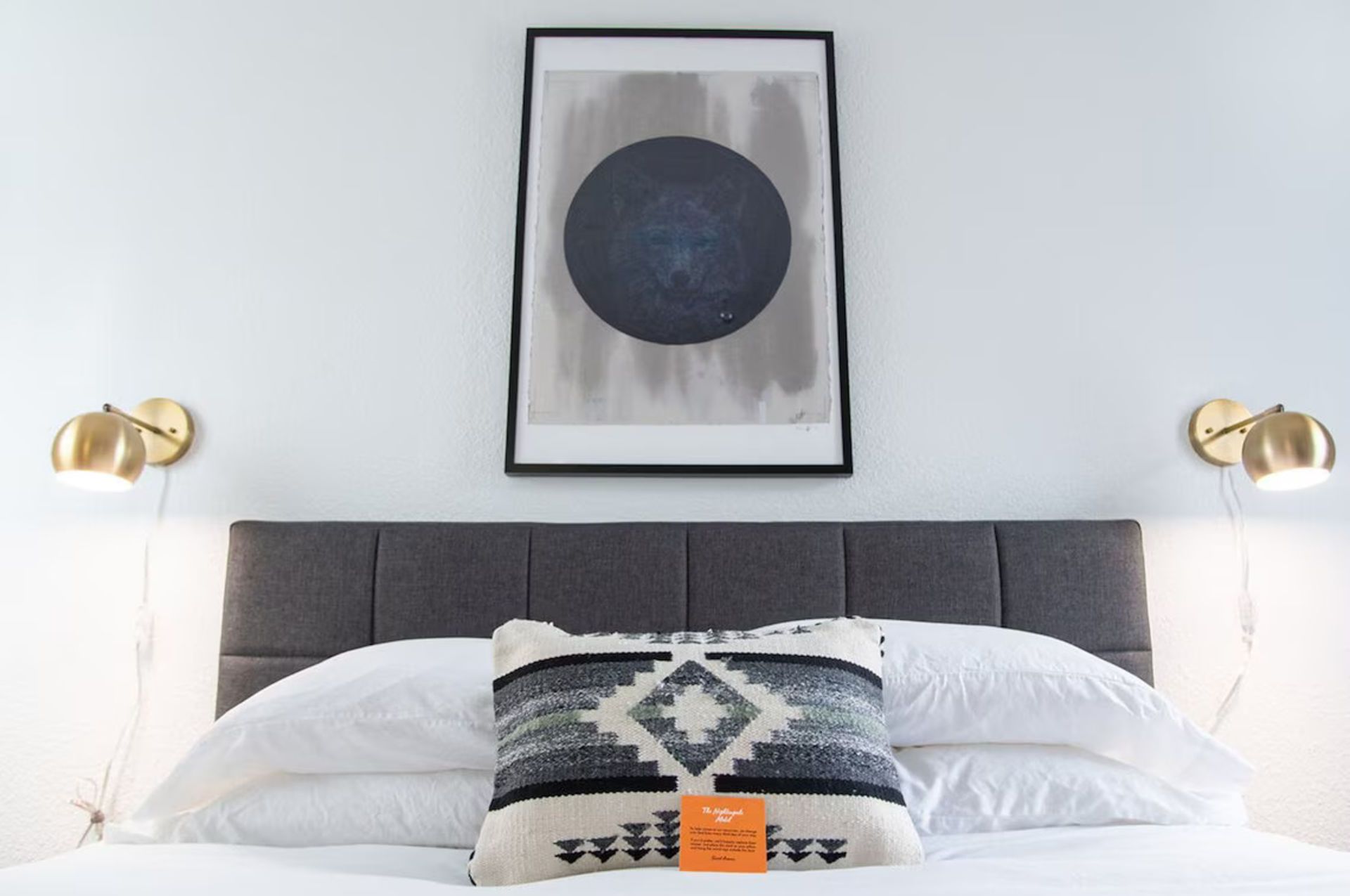 Bedroom with bed, headboard, art, gold sconces, and a patterned pillow.