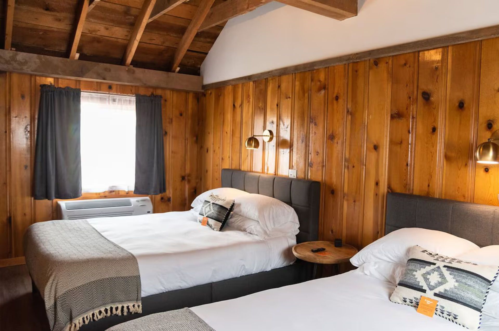 Two twin beds in a cabin room with wood paneling, small window, and two wall sconces.