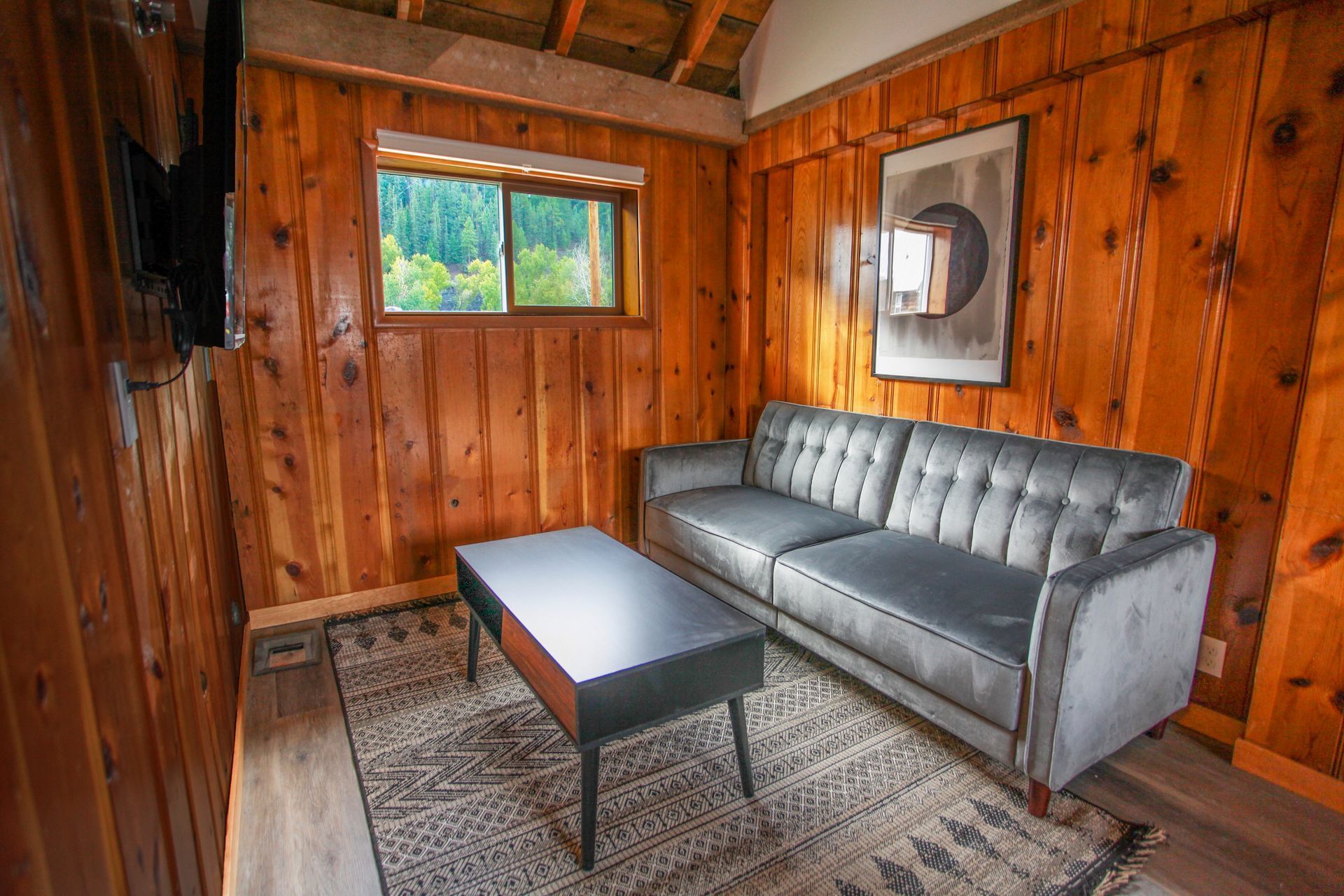 A cozy, wood-paneled living room with a gray couch, coffee table, and a window overlooking a forested area.