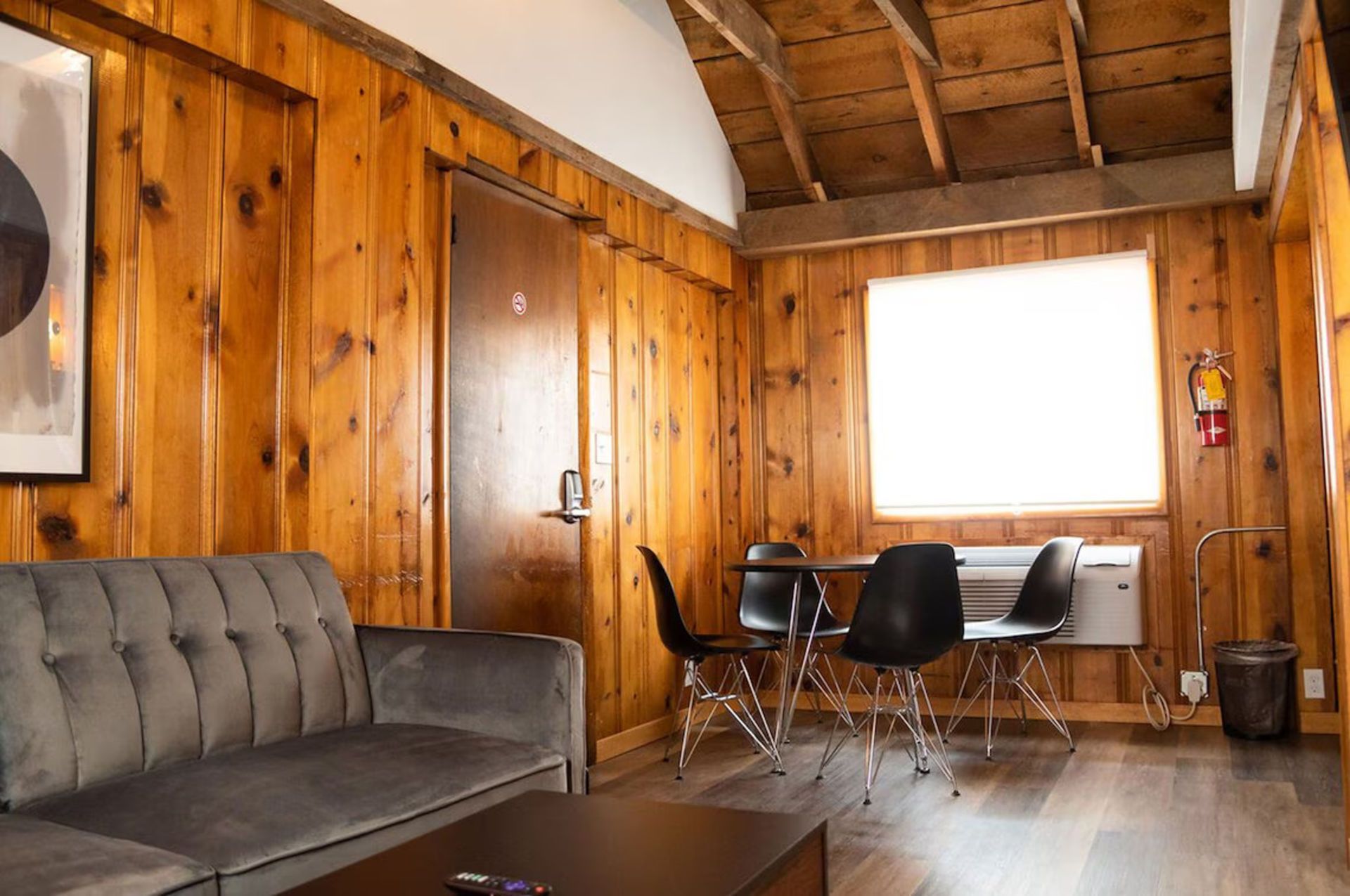 Cozy cabin interior with wood paneling, gray couch, small table and chairs, and window.
