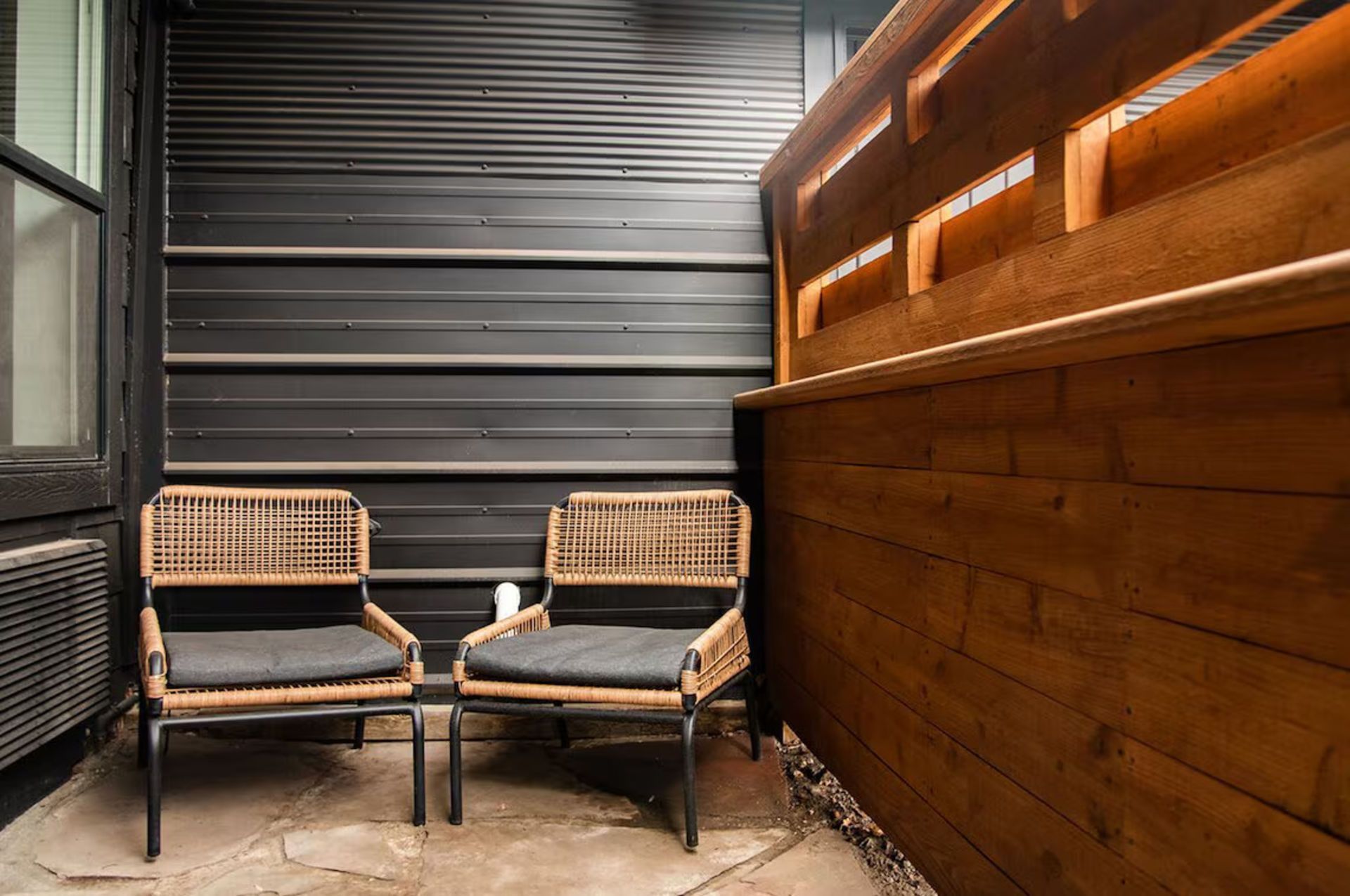Two wicker chairs with gray cushions on a small patio, next to a wooden fence.