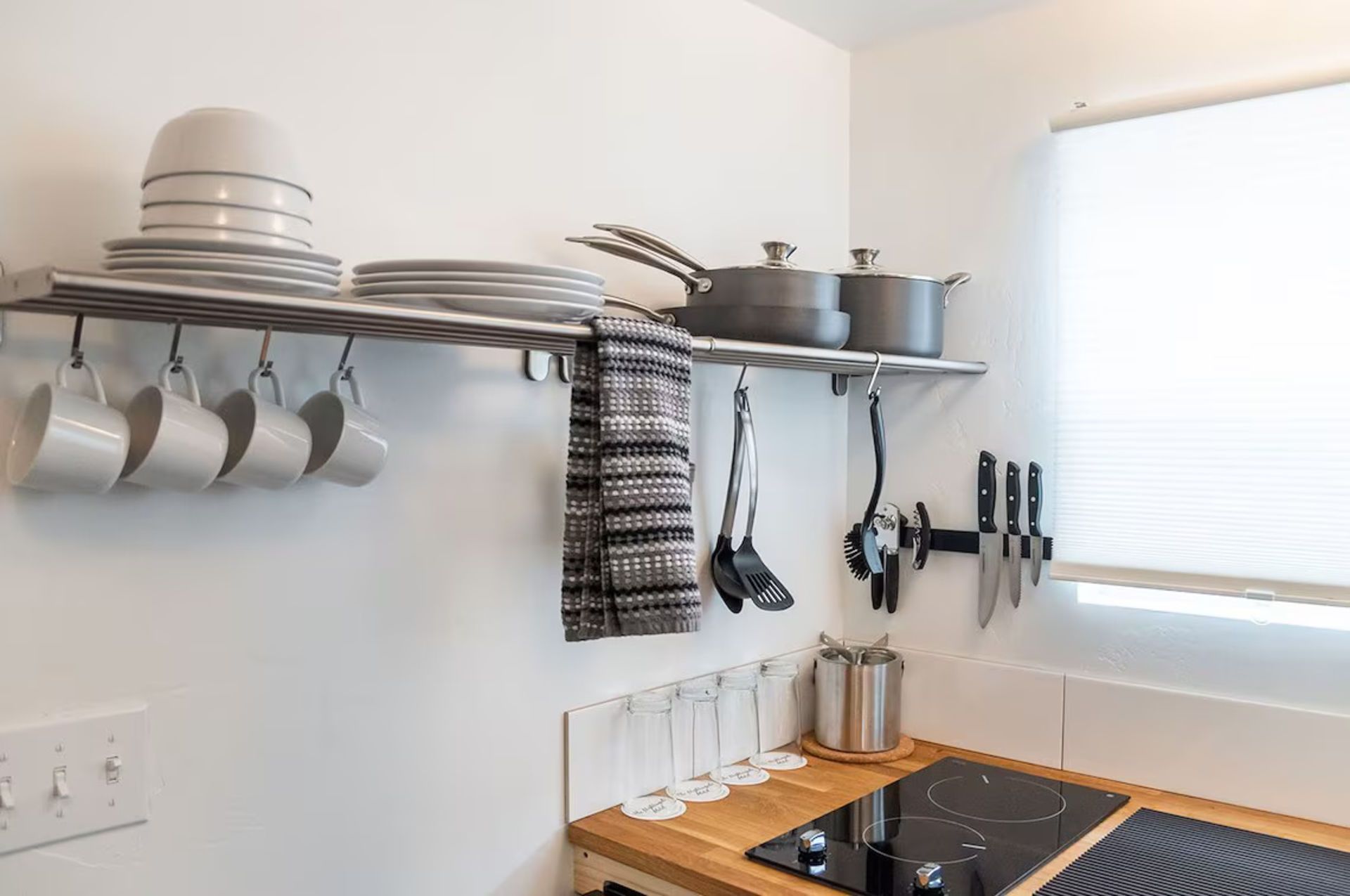 Kitchen shelf with dishes, mugs, pots, utensils, and knives near a stovetop.