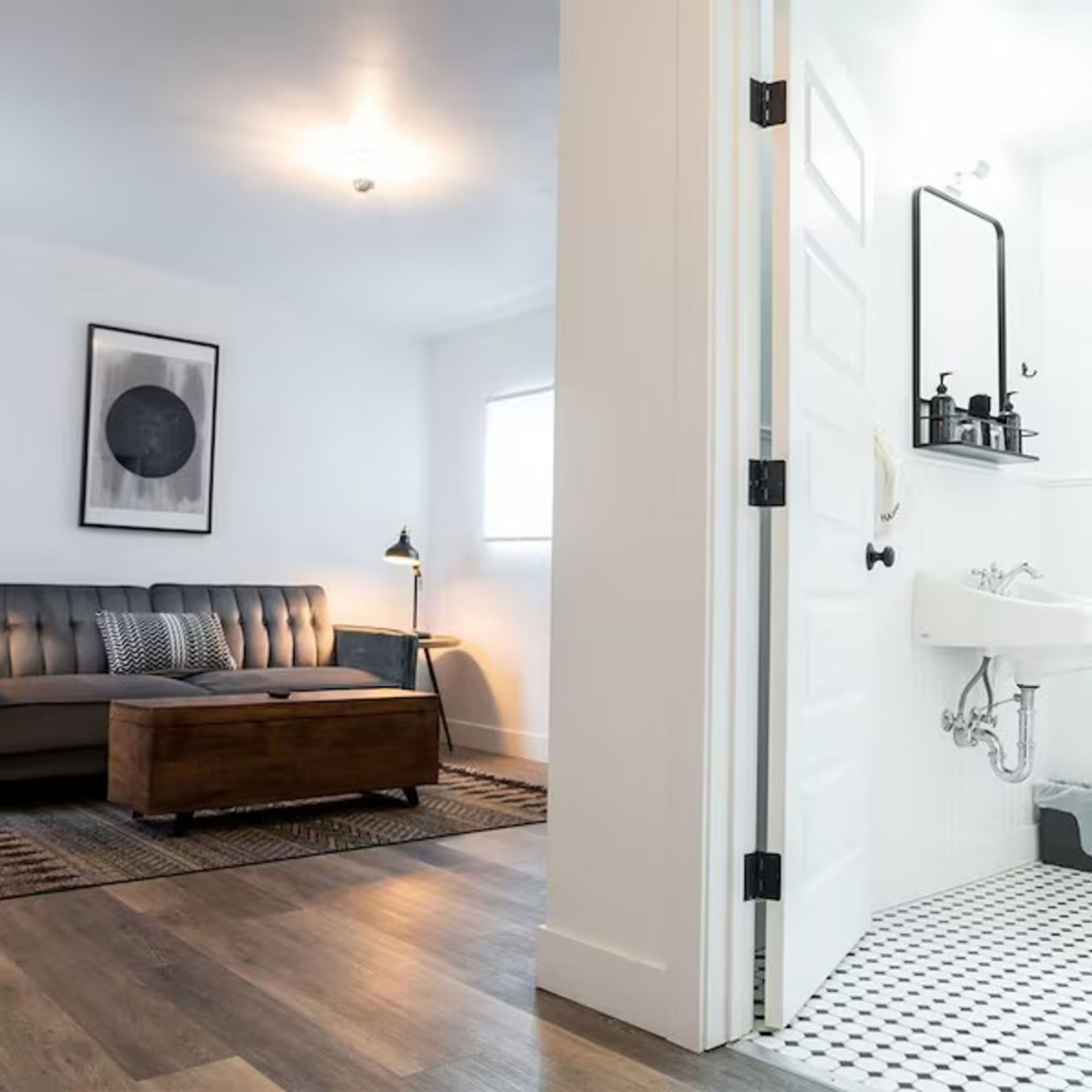 Living room with open doorway to a black and white tiled bathroom. Grey sofa, dark wood chest. White walls.