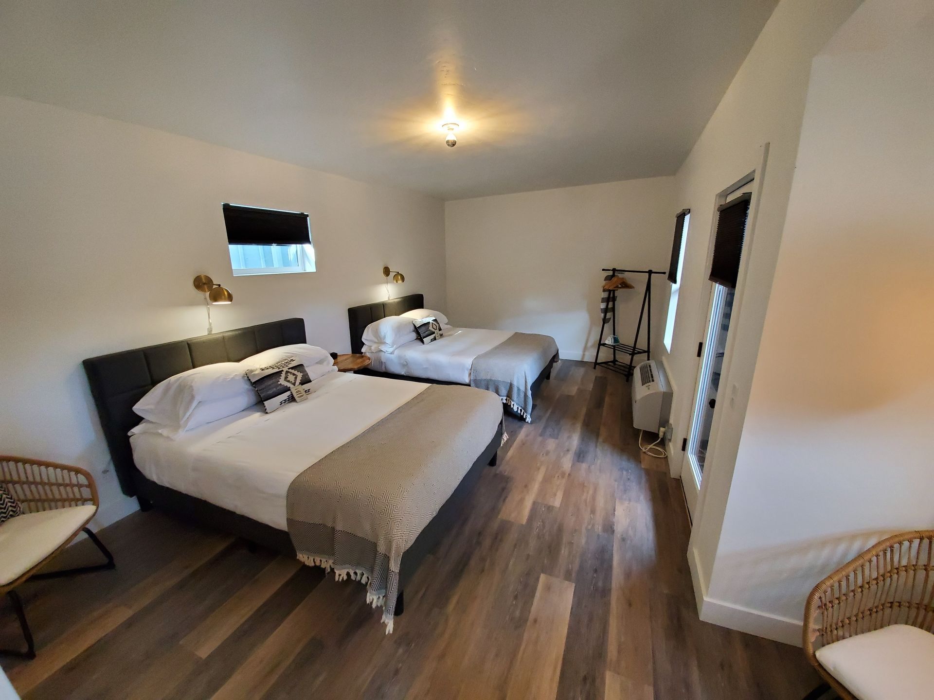 Two queen beds in a bright hotel room with wooden floors, two chairs, and a clothing rack.