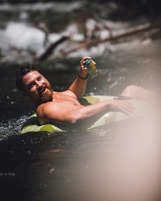Man floats on inner tube in river, smiling, holding a drink.