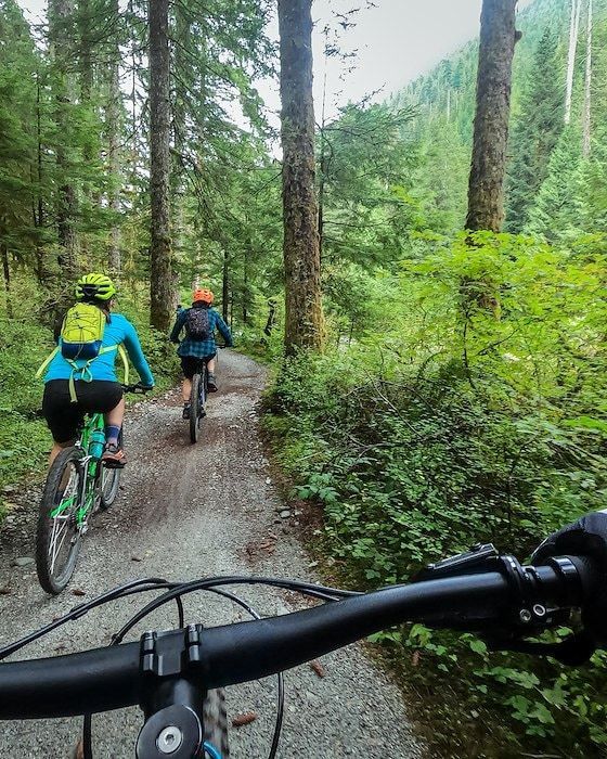 Two cyclists on a gravel path in a forest, following a trail. Focus is from the handlebars.