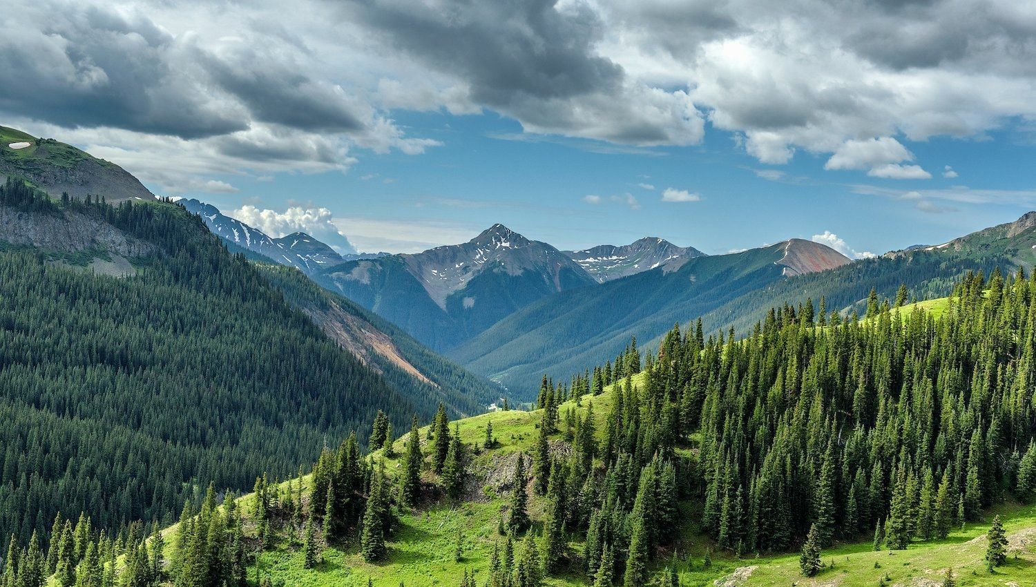 Mountain valley with green slopes, evergreen trees, and blue sky with clouds.