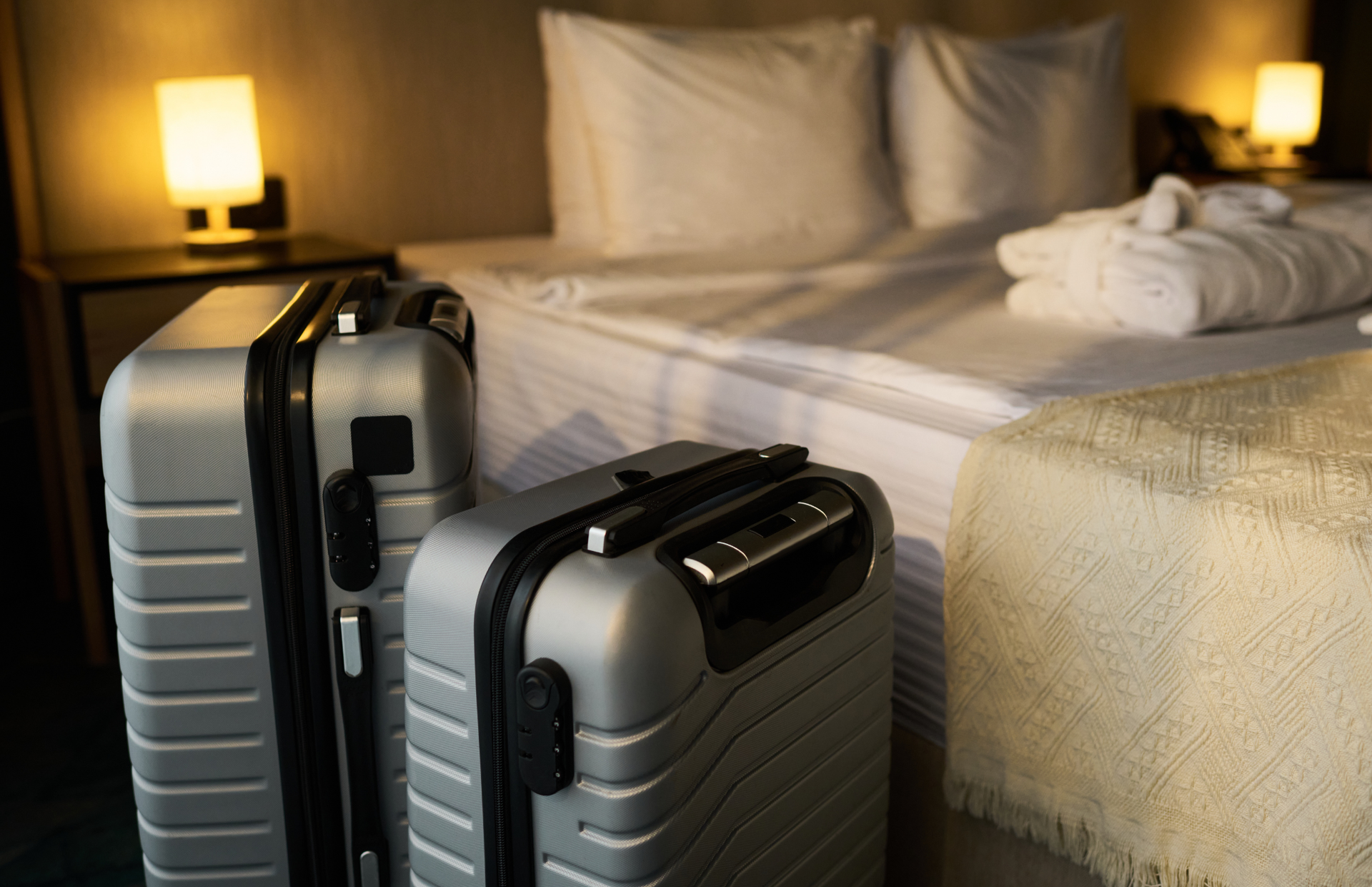 Two silver suitcases beside a neatly made bed in a softly lit hotel room